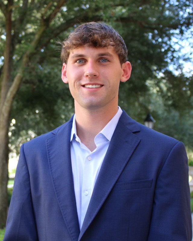 Portrait of Michael Niemann in a navy blazer, standing outdoors under oak trees